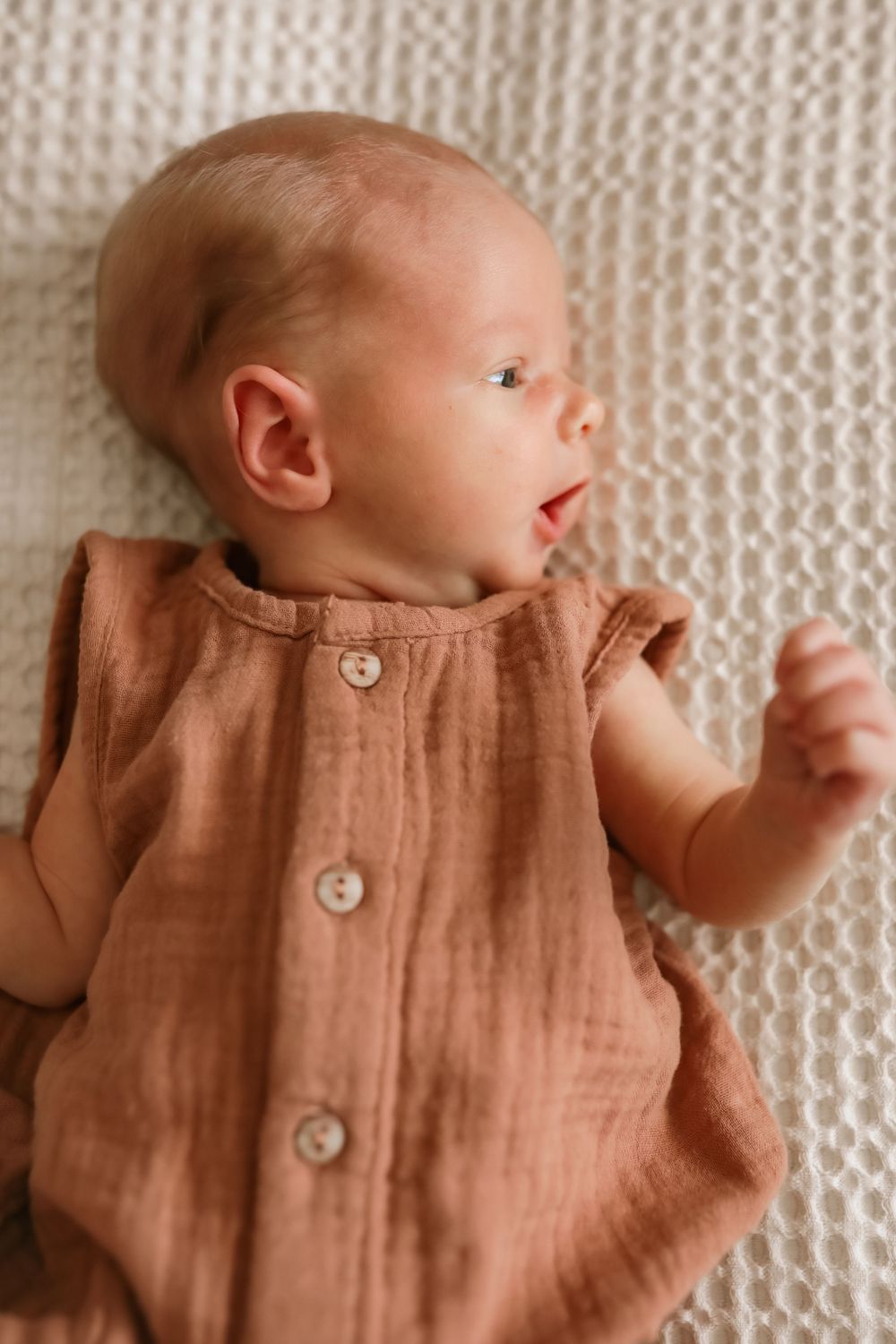 Newborn baby lying on white knit blanket wearing a rust brown muslin button-up romper.