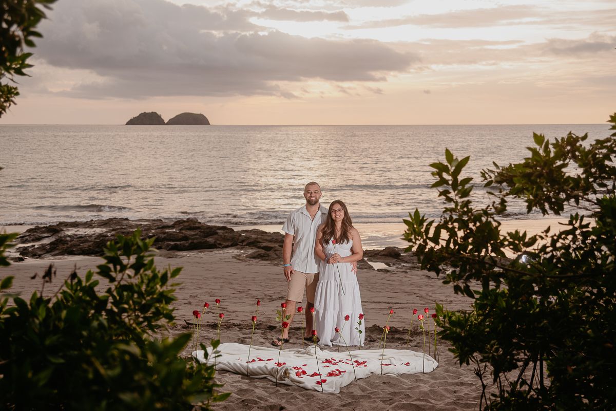 Couple embracing romantically on a sandy beach at sunset with calm ocean waves in the background.