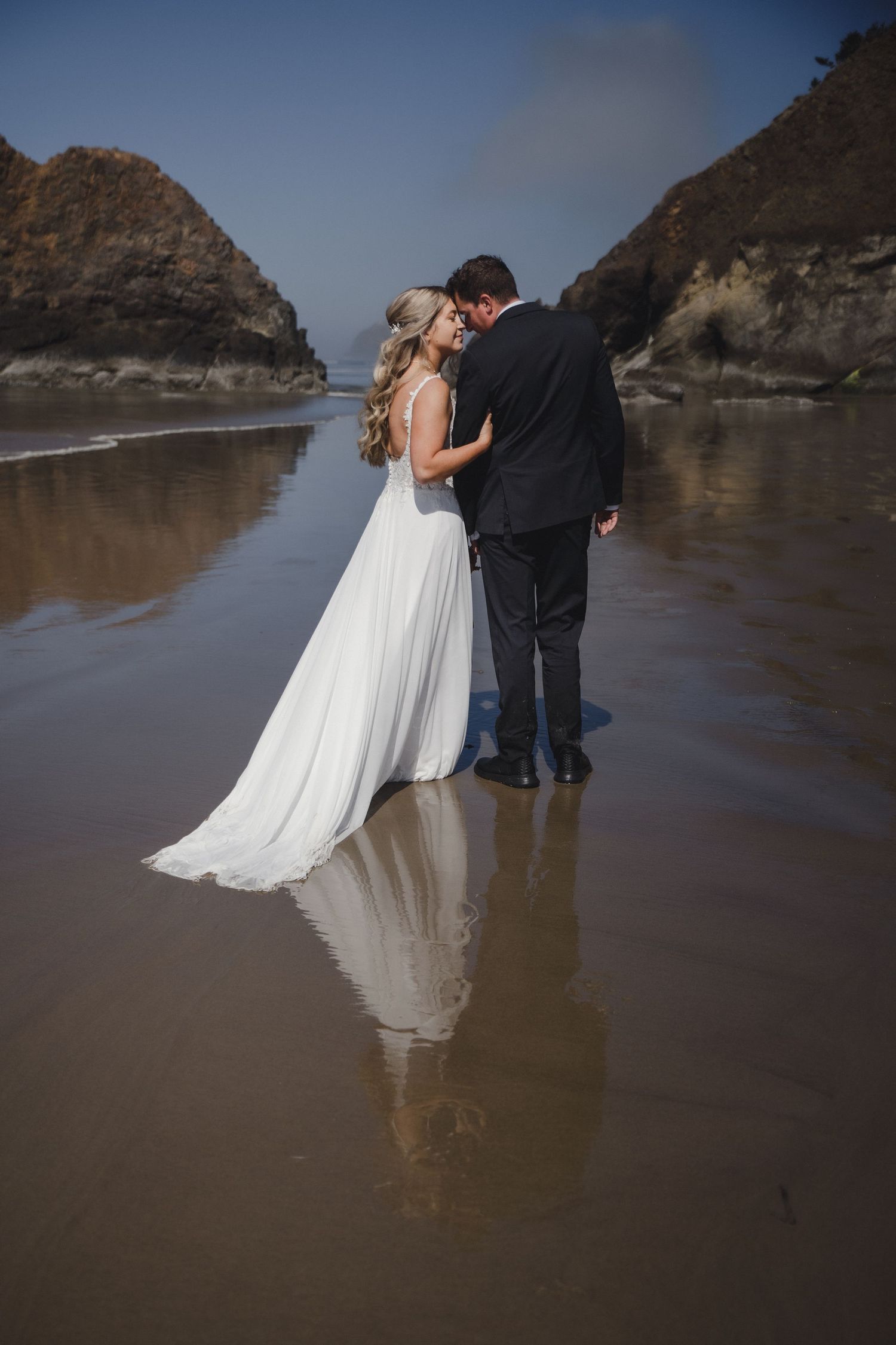 Bride and groom share a romantic moment on a reflective beach with rocky cliffs in the background.