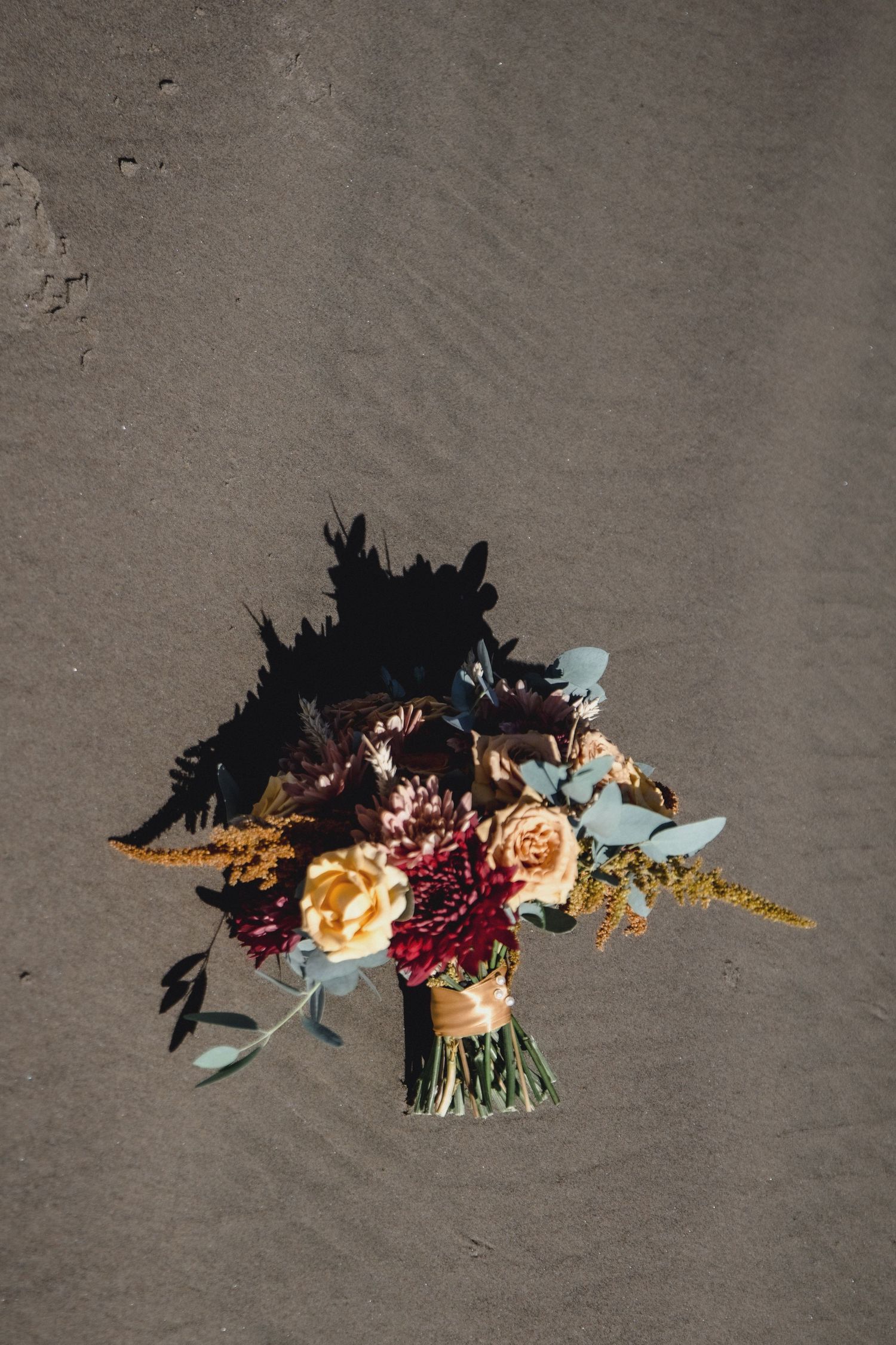 Bridal bouquet with burgundy, yellow, and blush roses on dark sand beach.