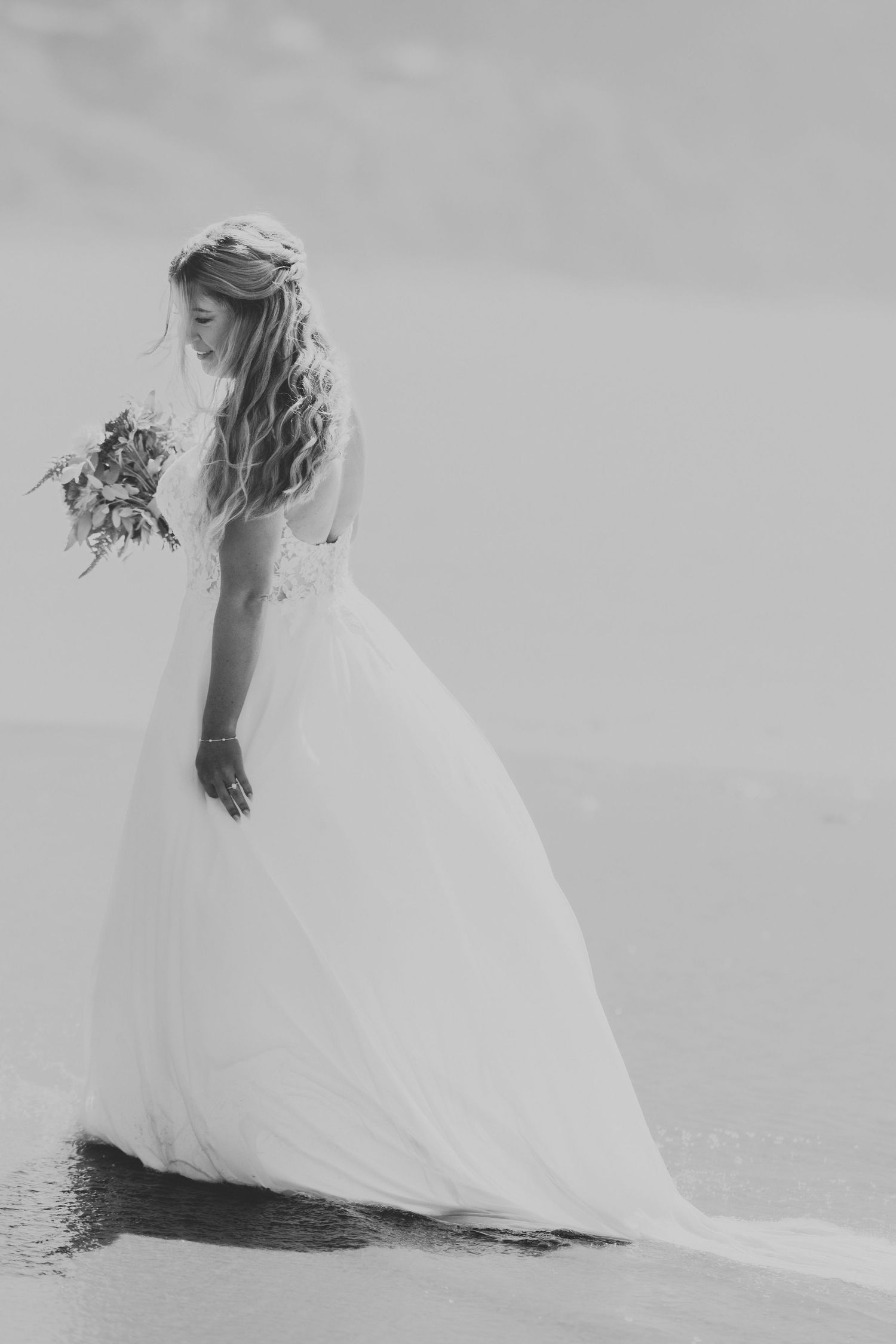 Bride in flowing white gown holding bouquet by ocean shore in black and white.