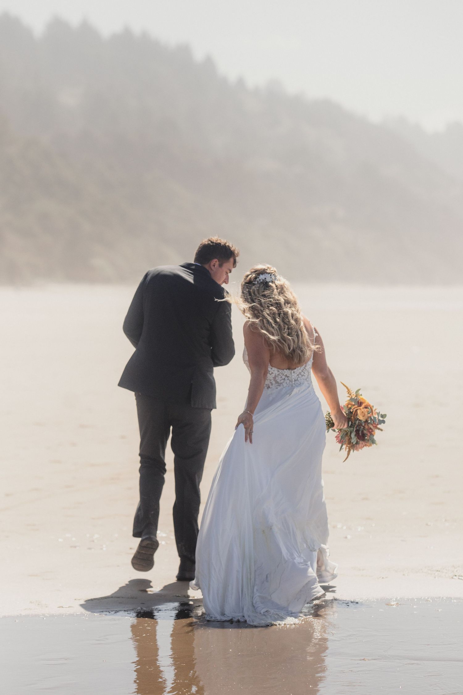 Bride and groom walking on a misty beach, bride holding a colorful bouquet in a white lace wedding dress.