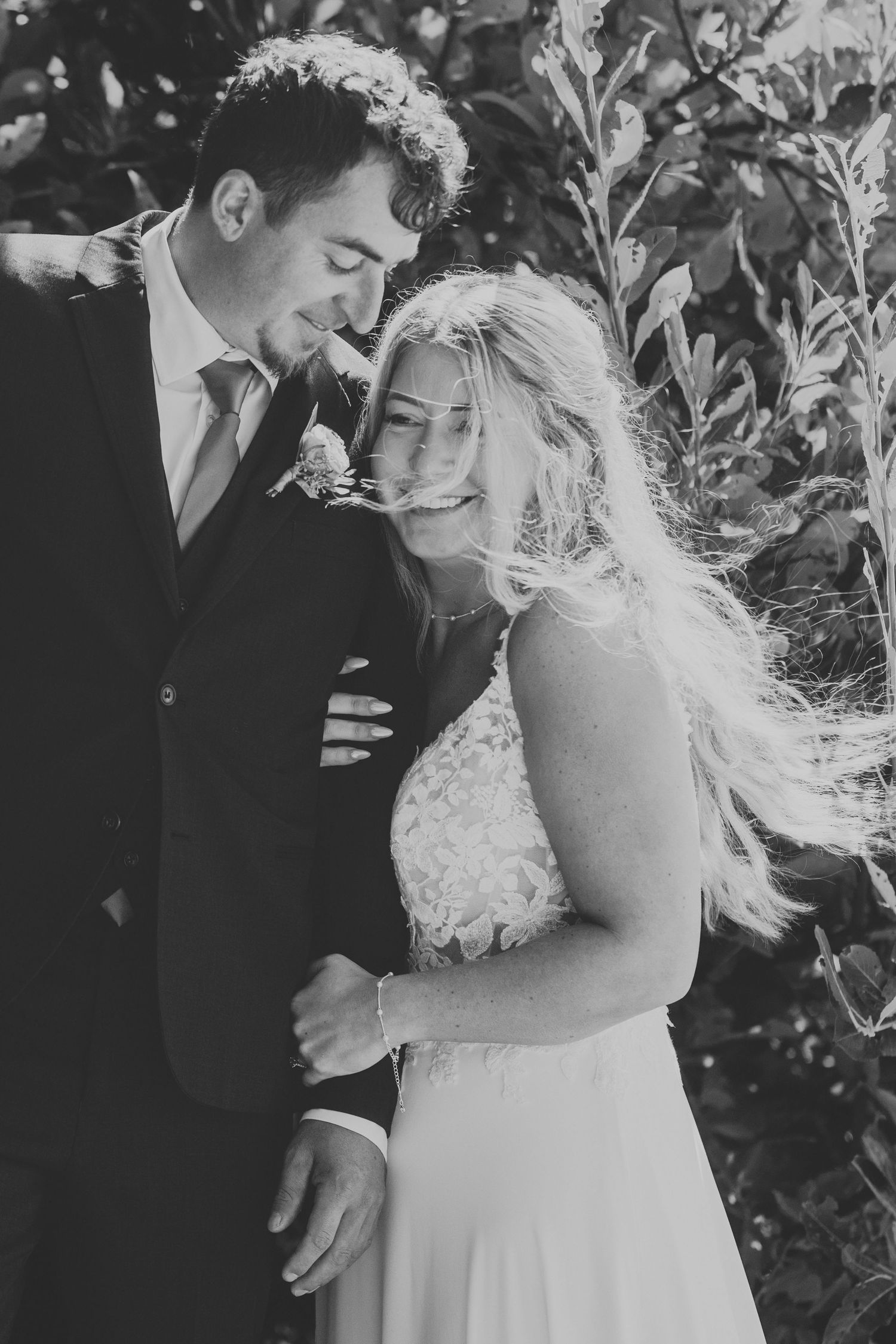 Black and white photo of smiling bride and groom embracing outdoors, bride in lace dress with flowing hair.