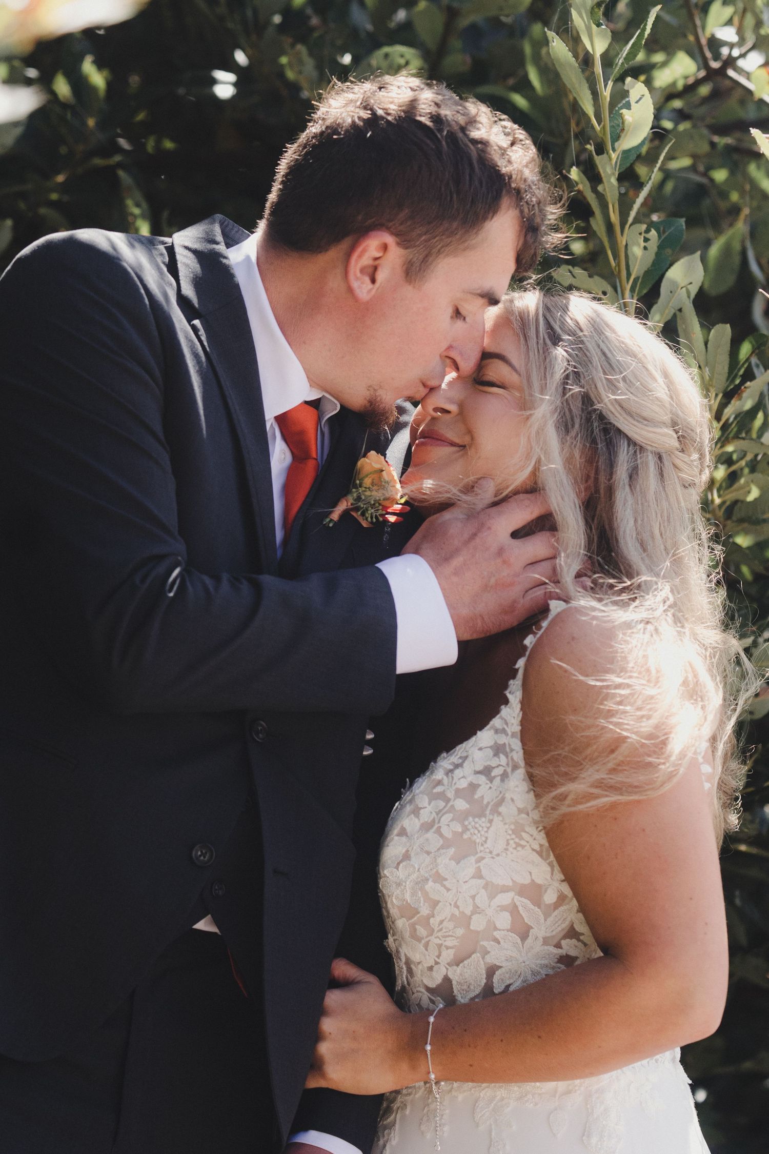 Bride and groom share a tender moment, touching foreheads outdoors, groom in dark suit with red tie.