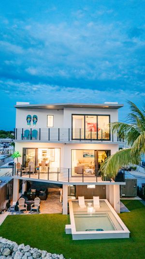 Modern two-story beachside villa with pool, glass railings, and illuminated interiors at dusk.