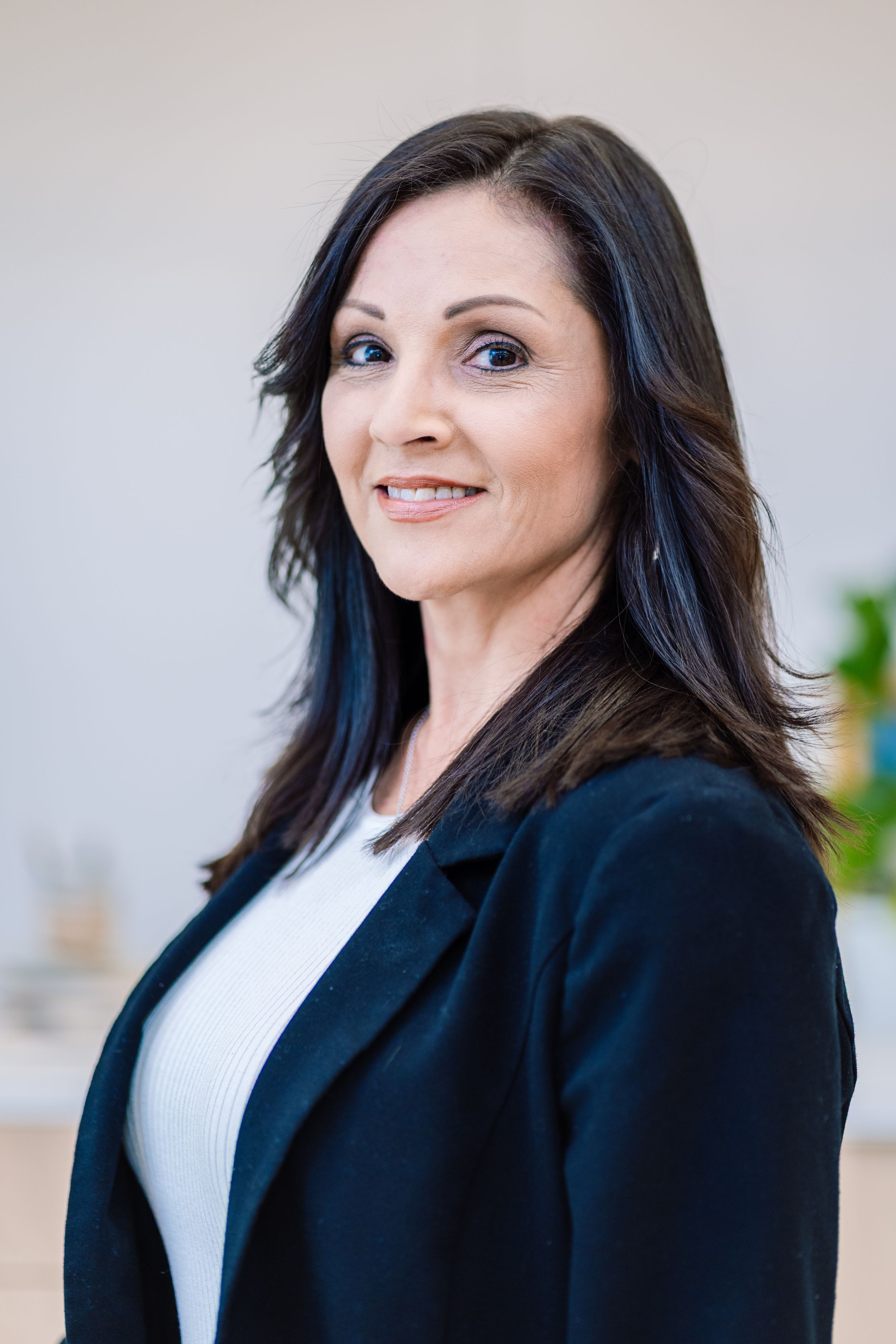 Professional woman in black blazer with long dark hair smiling confidently in a bright modern office setting.