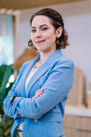 Professional woman in blue blazer standing with arms crossed, smiling confidently in a modern office setting.