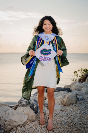 Smiling graduate in green cap and gown holds University of Florida Gators stole on rocky beach at sunset.