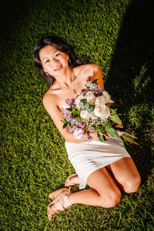 A smiling woman in a white dress sits on green grass holding a beautiful bouquet of white and purple flowers.