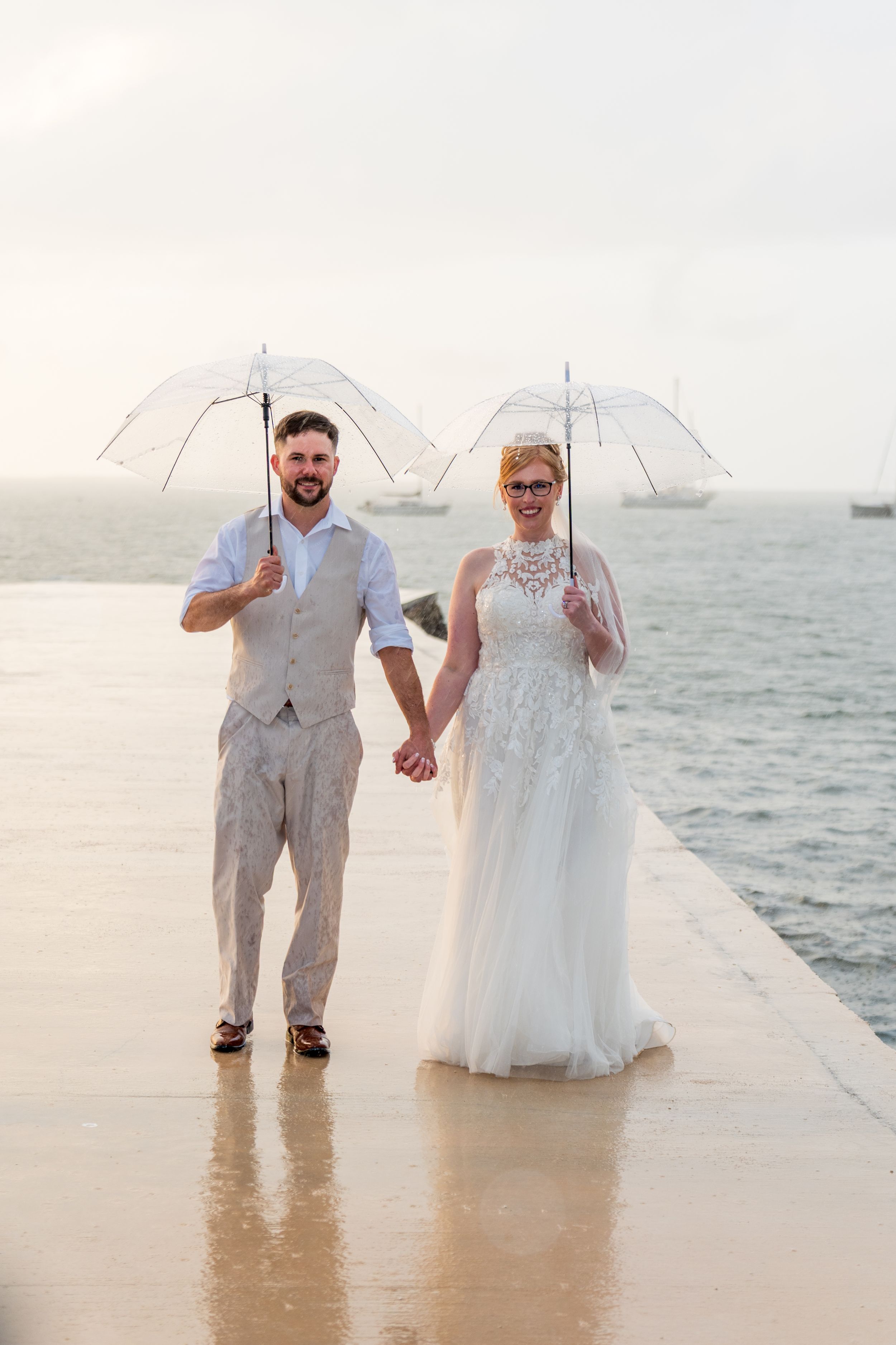 Bride and groom holding clear umbrellas on a wet pier by the ocean on their wedding day.