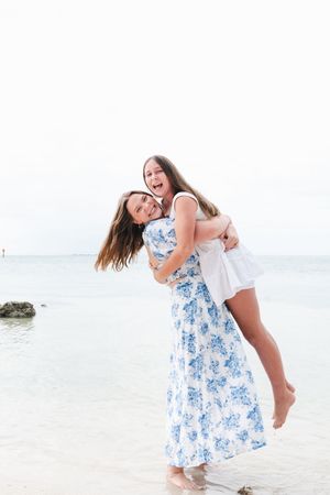 Two young women laughing and embracing on a tropical beach, one wearing a blue floral maxi dress giving the other a piggyback ride.