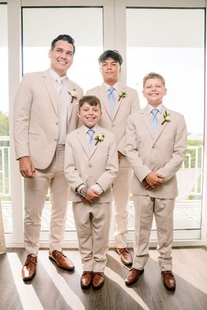 Four males in matching tan suits with boutonnieres and blue ties pose together at a wedding ceremony.