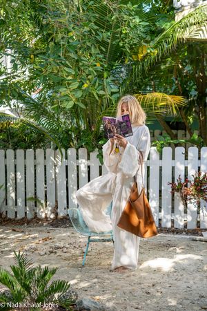 Blonde woman in white linen outfit reading a book on a blue chair in a tropical garden with white picket fence.