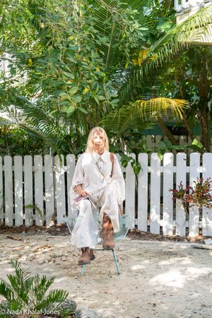 Blonde woman in flowing white outfit standing by a blue chair in a sunny tropical garden with white picket fence.