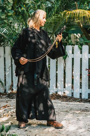 Blonde woman in black outfit holding a leash outdoors near white picket fence and tropical plants.