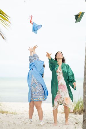 Two women in colorful dresses tossing hats in the air on a tropical beach near a palm tree.
