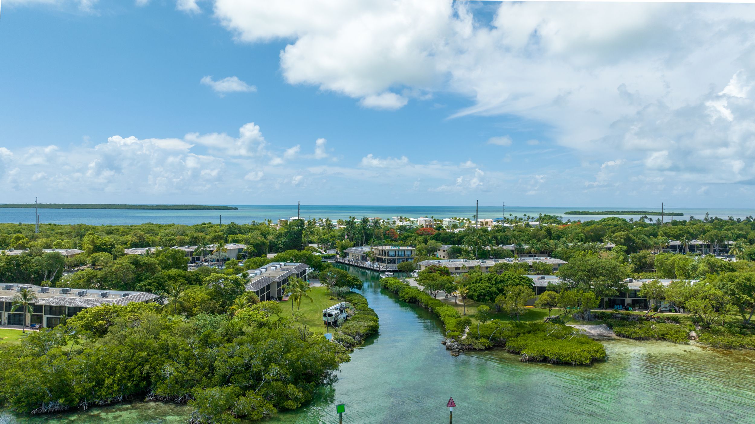 Aerial view of a tropical island canal surrounded by lush green mangroves and waterfront homes under a bright blue sky.