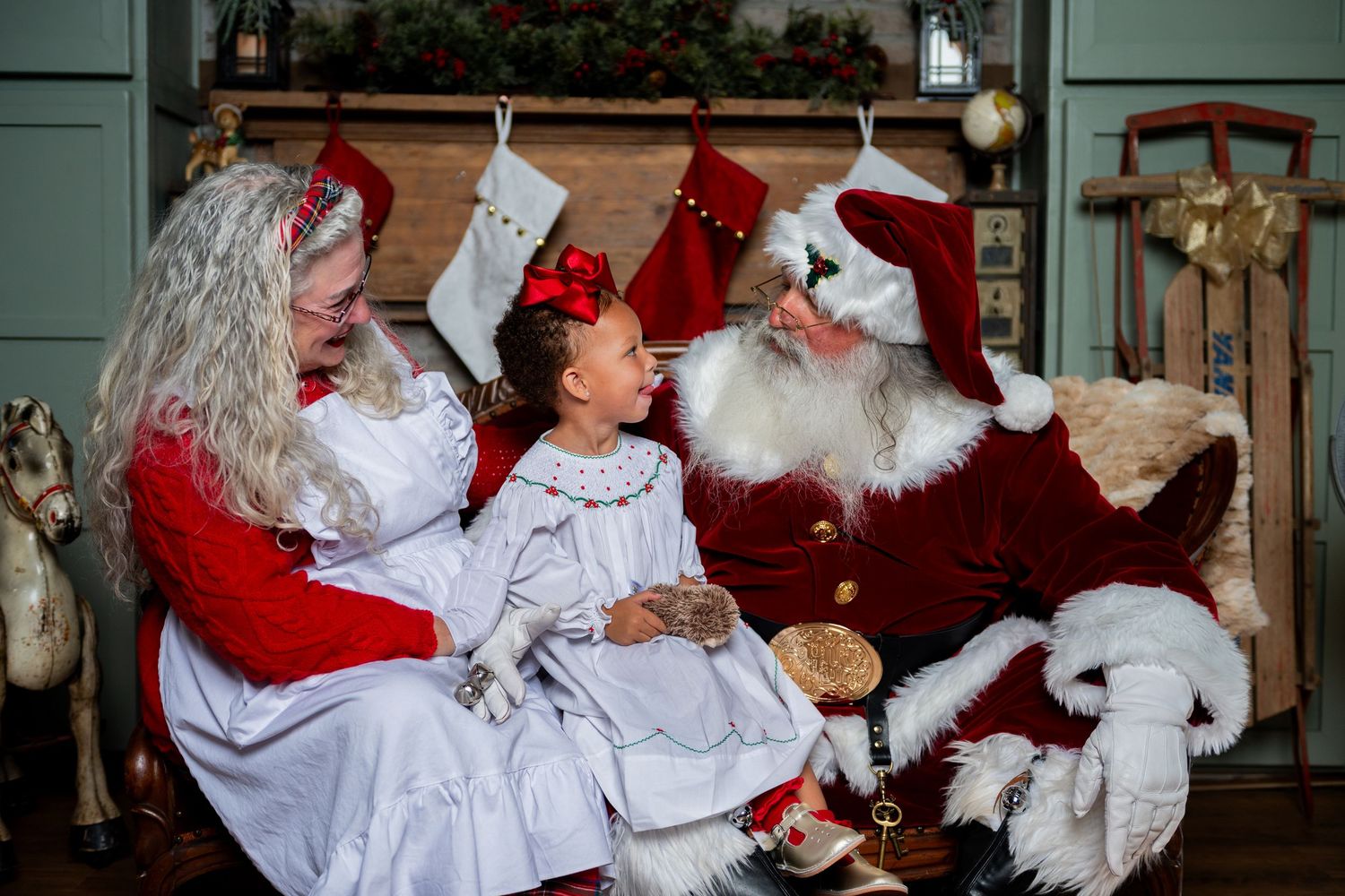 A young girl sits between Santa Claus and Mrs. Claus in a festive holiday setting with stockings and a sleigh.