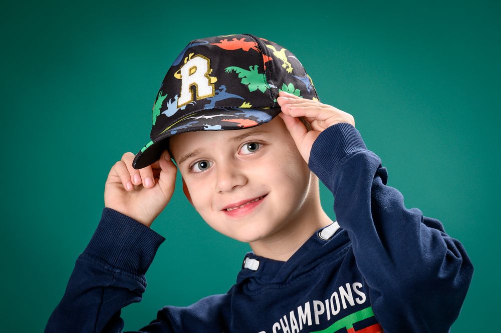 Child in sports cap and navy jacket poses against teal studio backdrop.