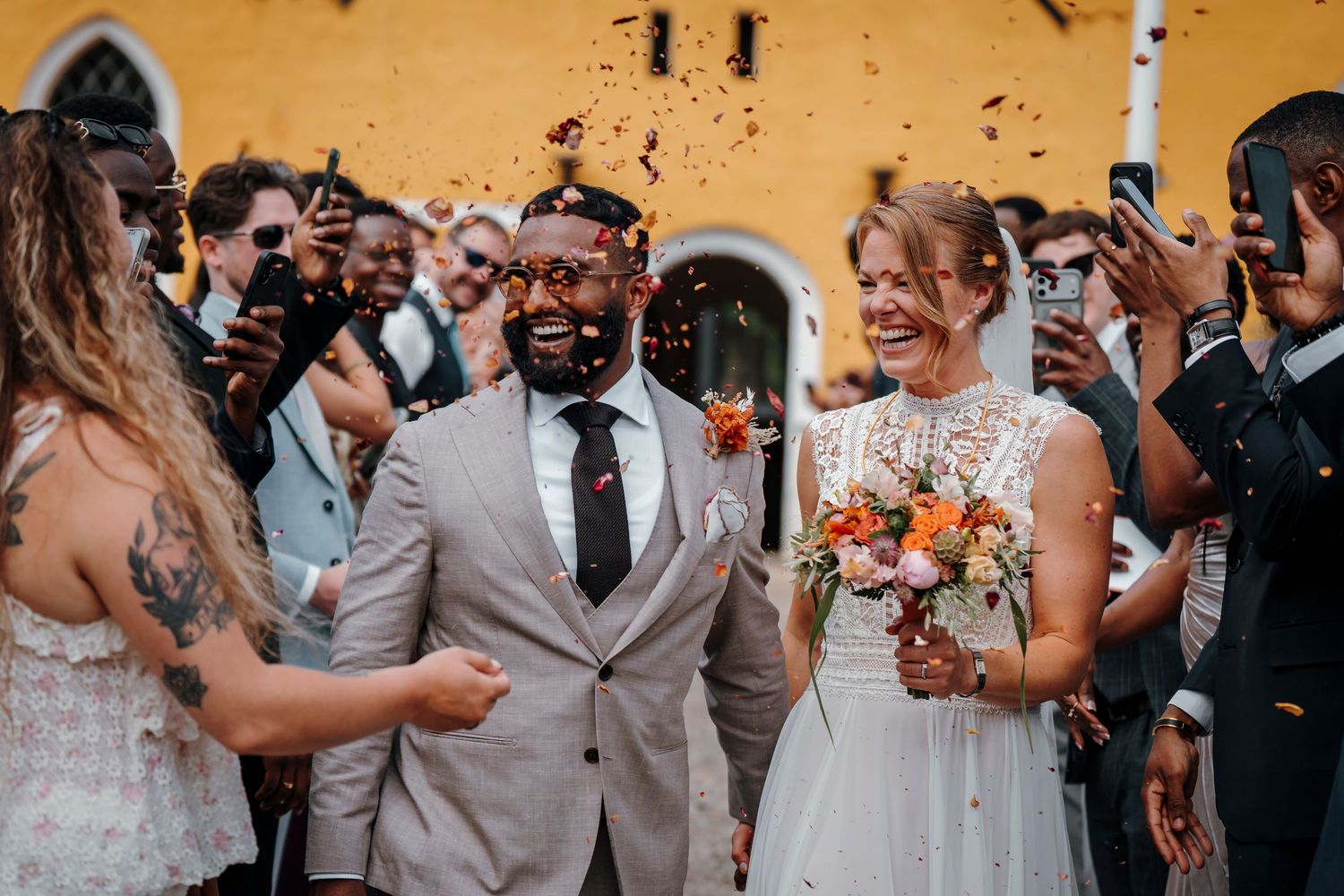 Joyful newlywed couple walks through confetti shower surrounded by cheering guests at outdoor wedding ceremony.