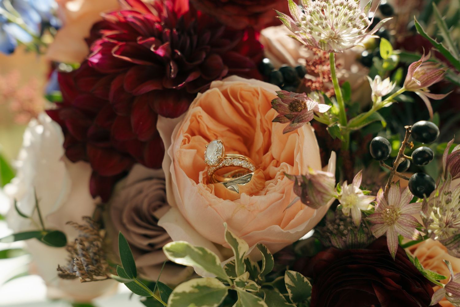Wedding rings nestled in a bridal bouquet with peach garden roses, burgundy dahlias, and lush greenery.