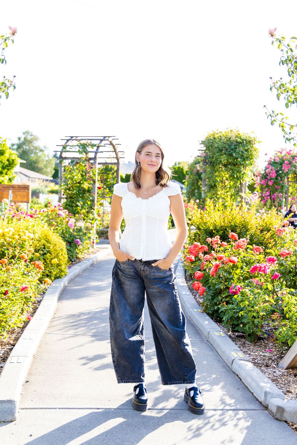 Young woman in white top and wide-leg jeans poses on garden path surrounded by colorful rose bushes.