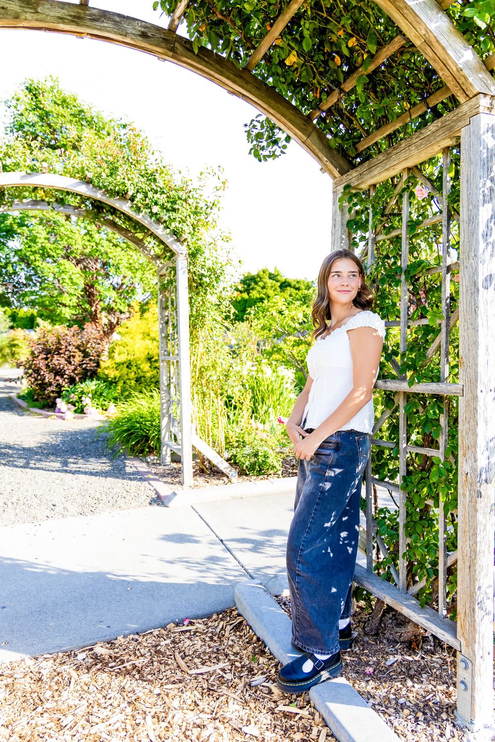 Young woman in white top and jeans leaning against a wooden garden arbor surrounded by lush green plants.