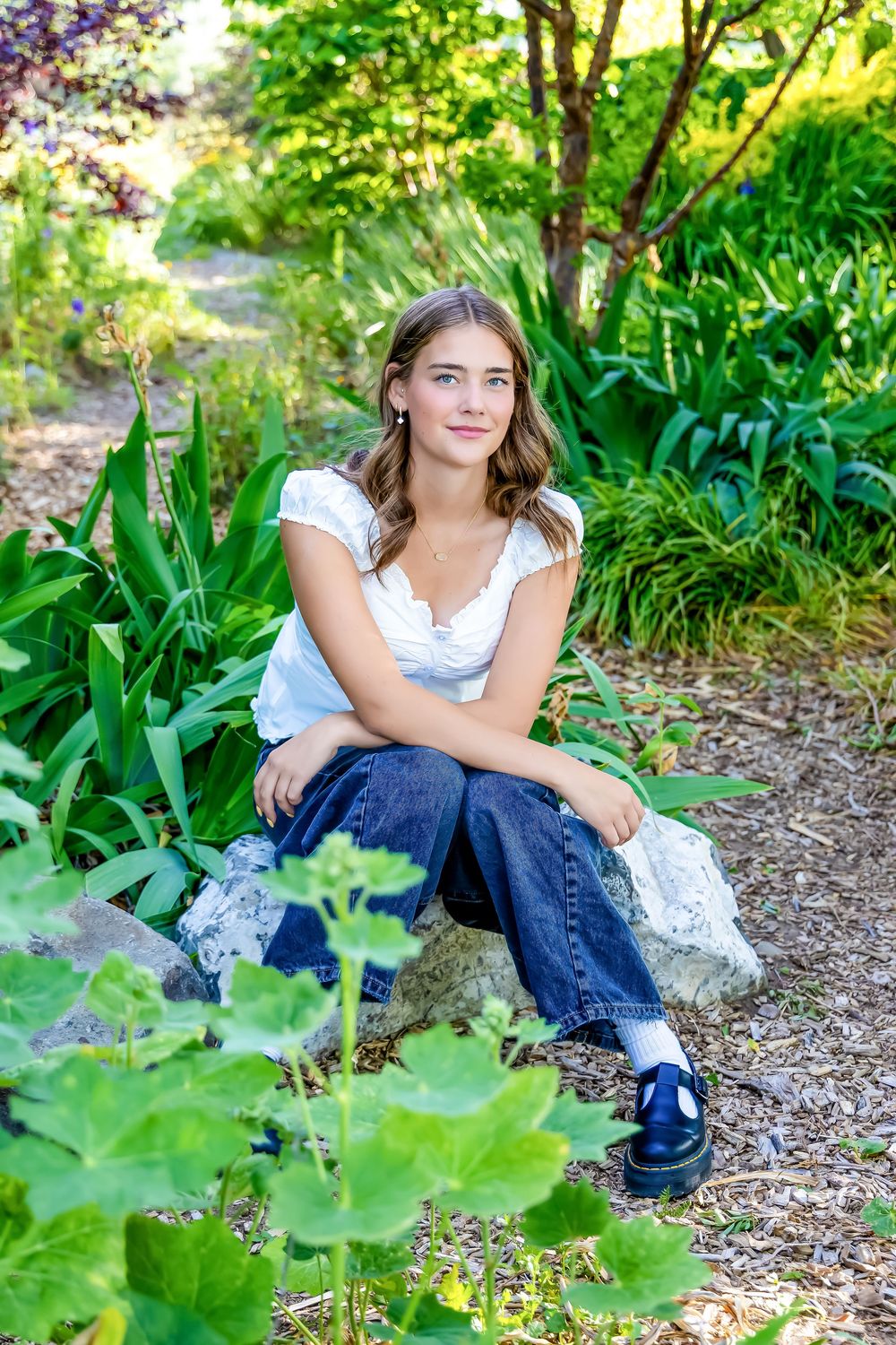 Young woman in white top and jeans sits on a rock surrounded by lush green garden plants.