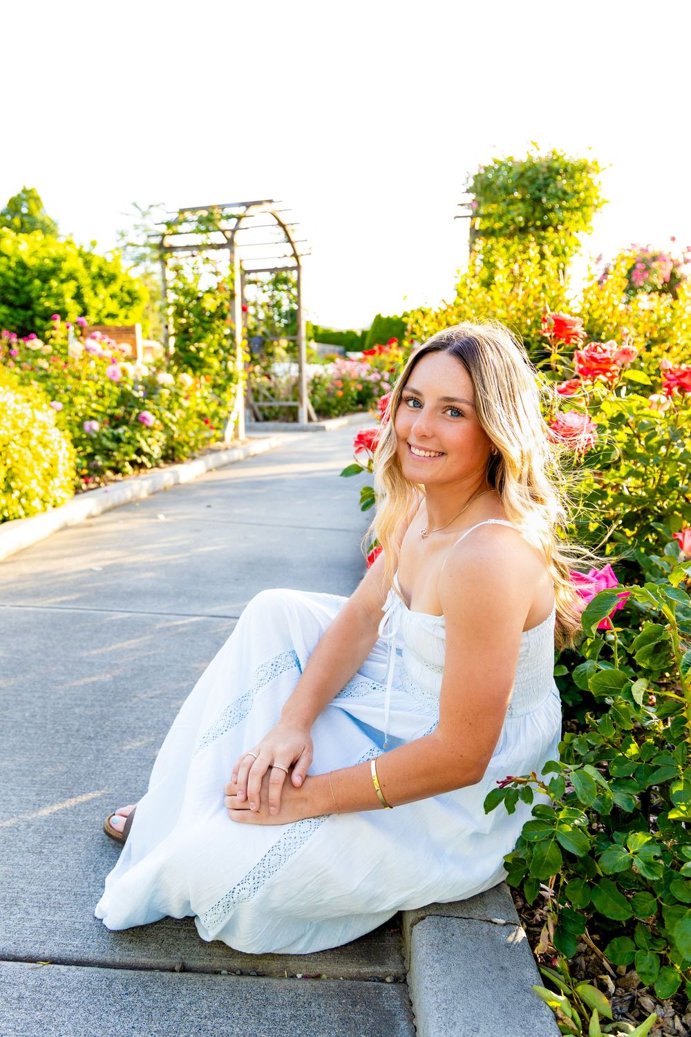 Young blonde woman in white dress sitting on garden path surrounded by colorful roses on sunny day.