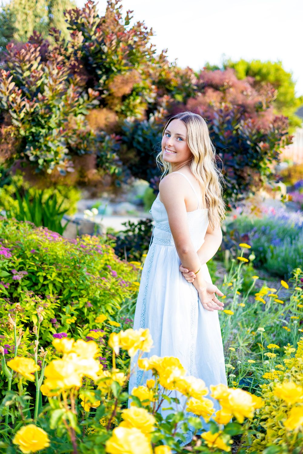 Young woman in white dress smiling among colorful garden flowers including yellow roses and lush greenery.