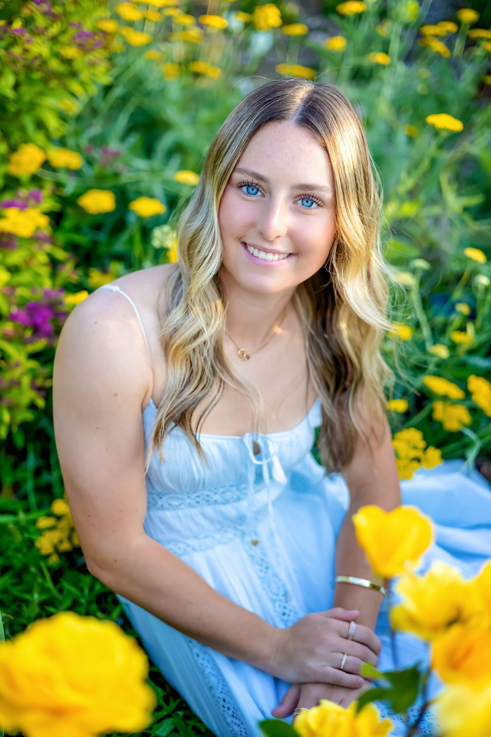 Young blonde woman with blue eyes smiles among vibrant yellow flowers in a garden, wearing a white dress.
