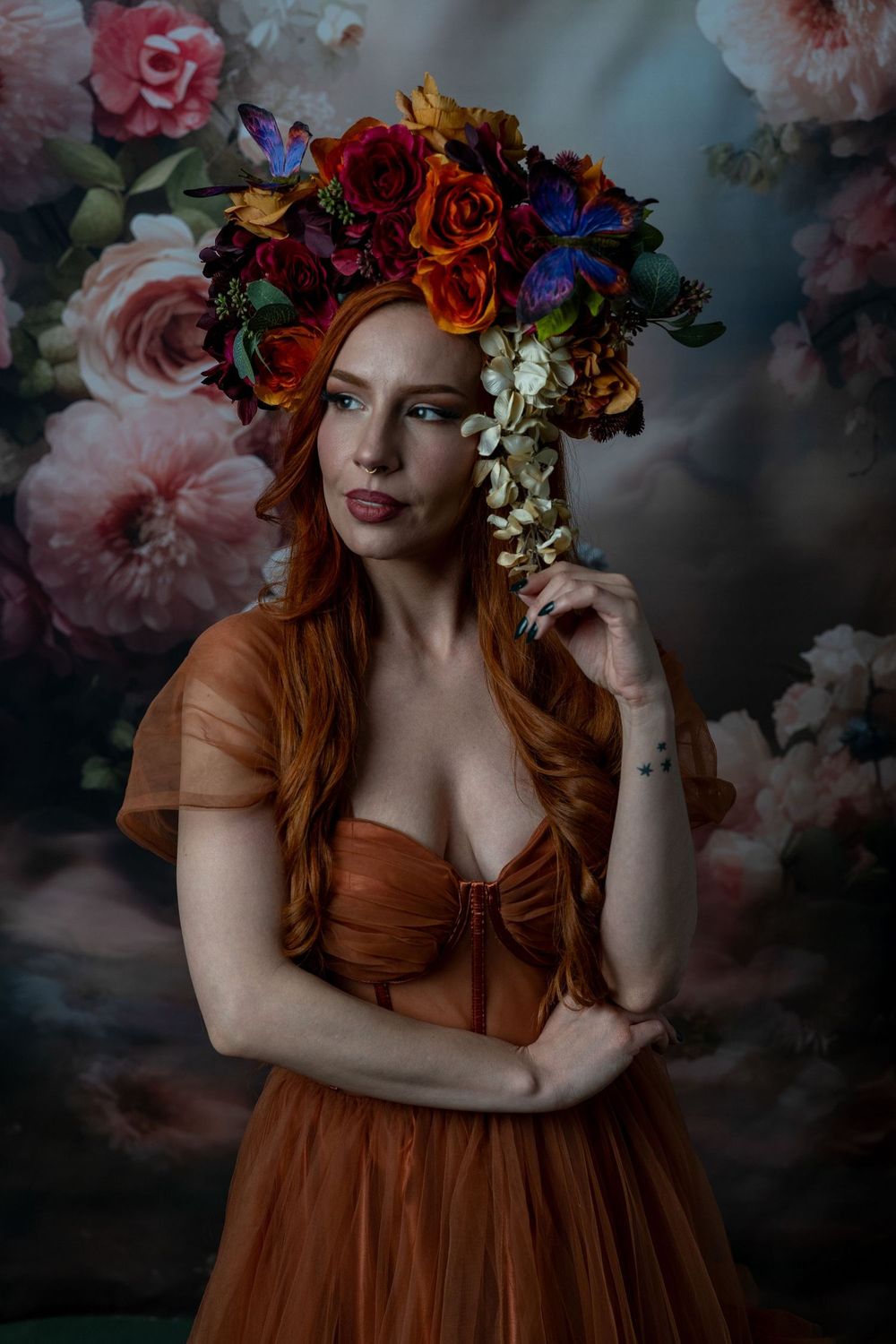 A woman with long red hair wearing a colorful floral crown and rust orange tulle gown poses before a floral backdrop.