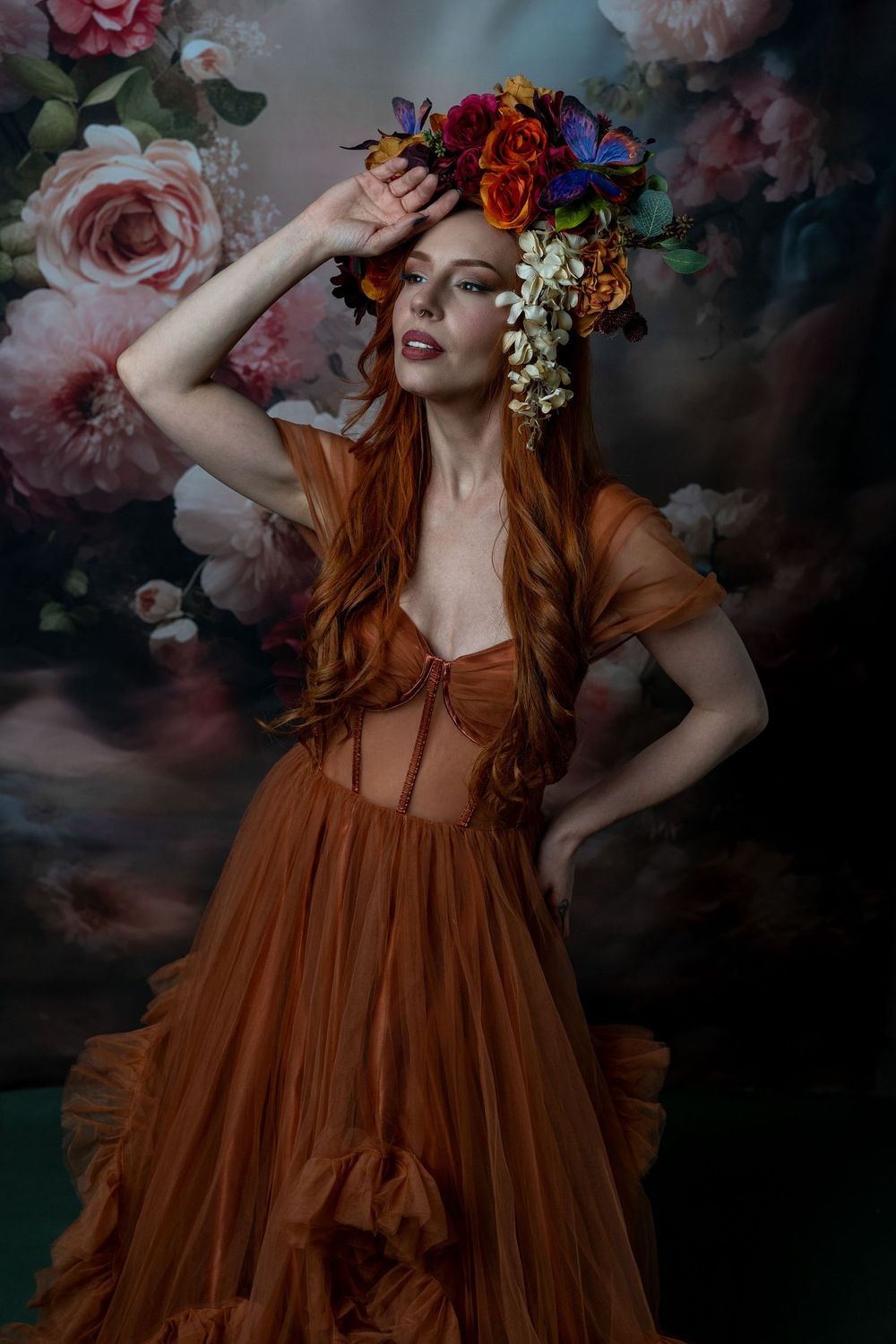Woman in rust tulle gown and floral crown poses against dreamy floral backdrop with long auburn hair.