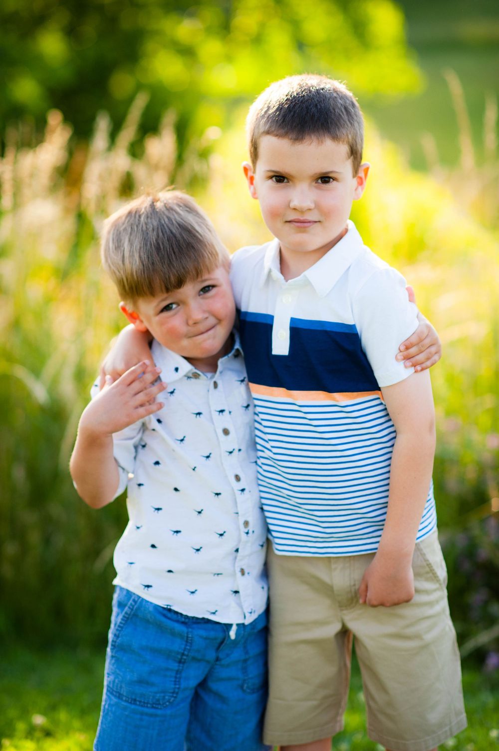 Two young boys hugging outdoors in a sunny green field, wearing casual summer clothes and smiling.