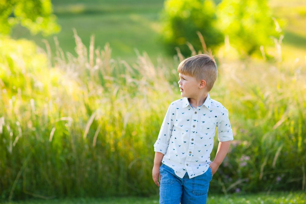 Young boy in white shirt and blue jeans standing outdoors in a sunny green field.