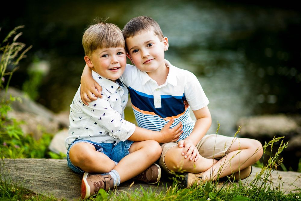 Two young boys sitting together outdoors near a creek, smiling and hugging in a sunny green setting.