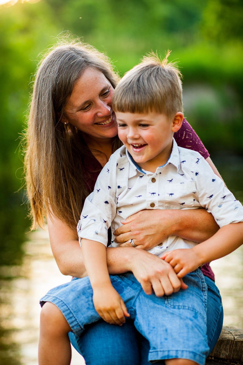 A smiling mother hugs her laughing young son outdoors near a pond with lush green trees in the background.