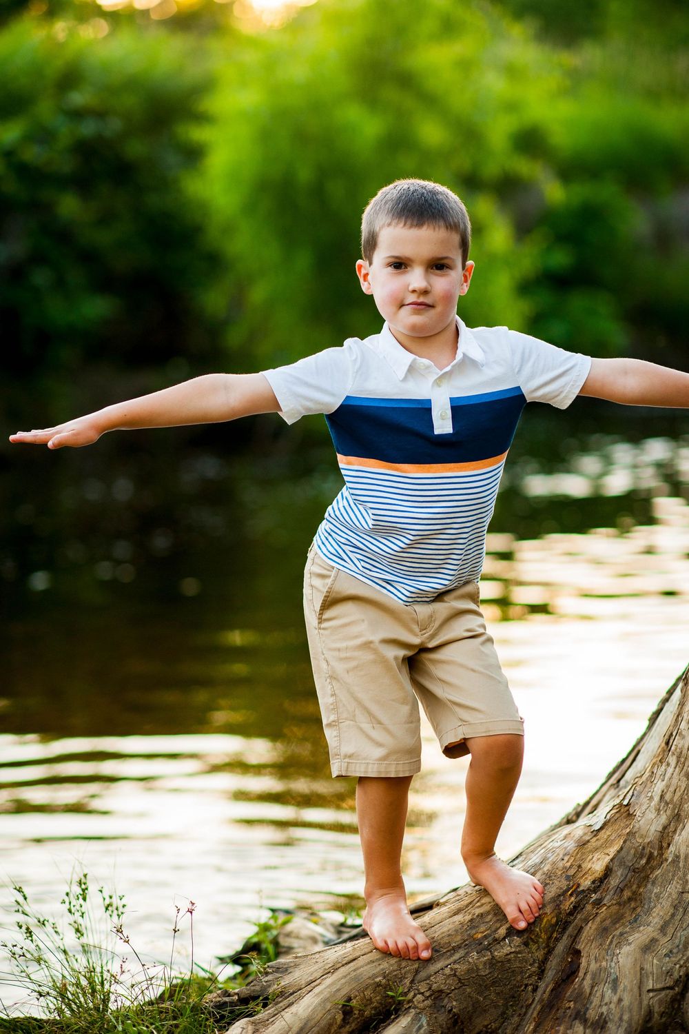 A young boy balancing on a log with arms outstretched near a sunlit river surrounded by lush green trees.