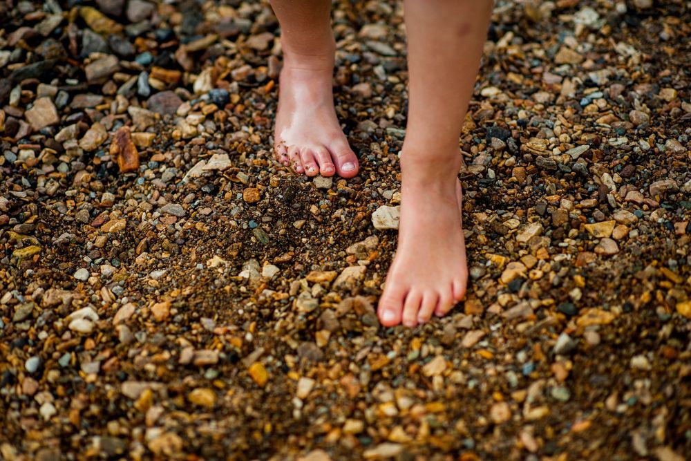 Bare feet with red toenails standing on a rocky pebble beach shoreline.