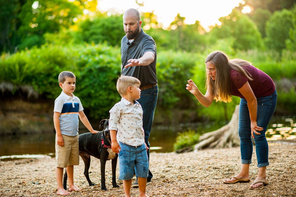 Family with two boys and a black dog enjoying a sunny outdoor moment near a river surrounded by lush greenery.