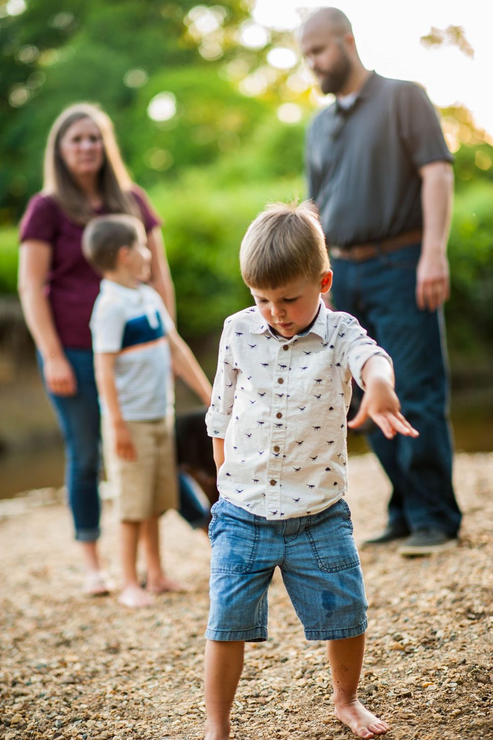 A young boy skips stones by a creek while his family watches in the background on a sunny day.