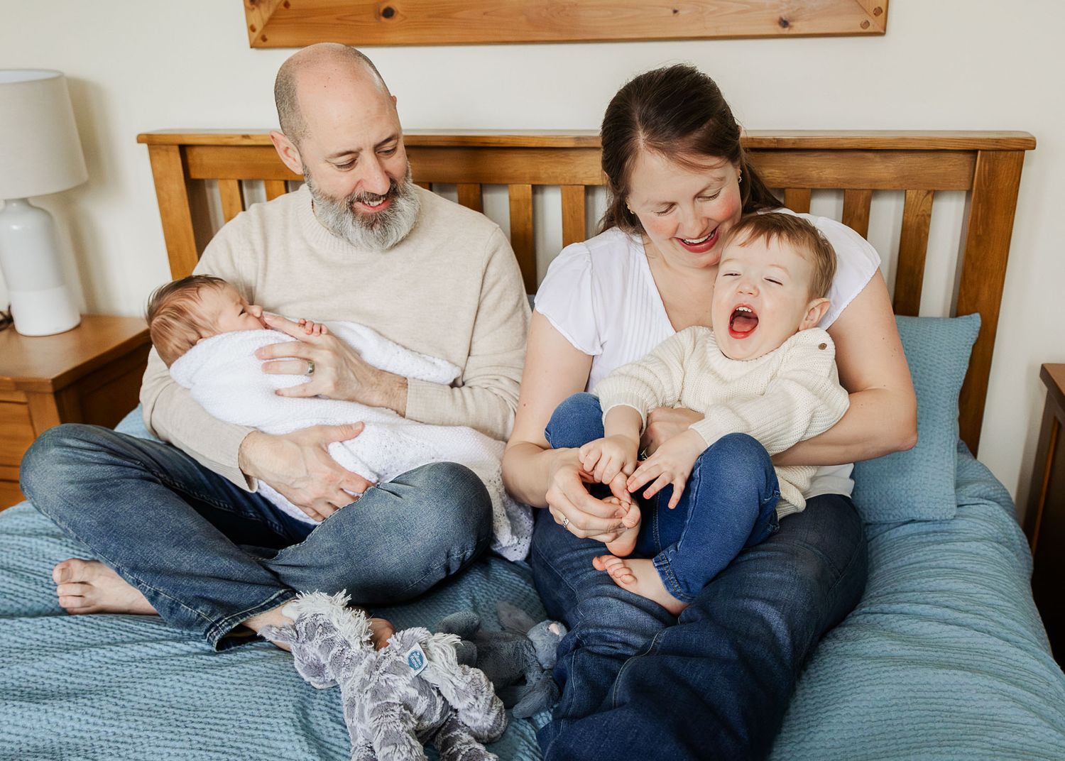 A newborn lies in dad's arms as his older brother is tickled by mum on their bed at home in Bristol during a relaxed family photoshoot
