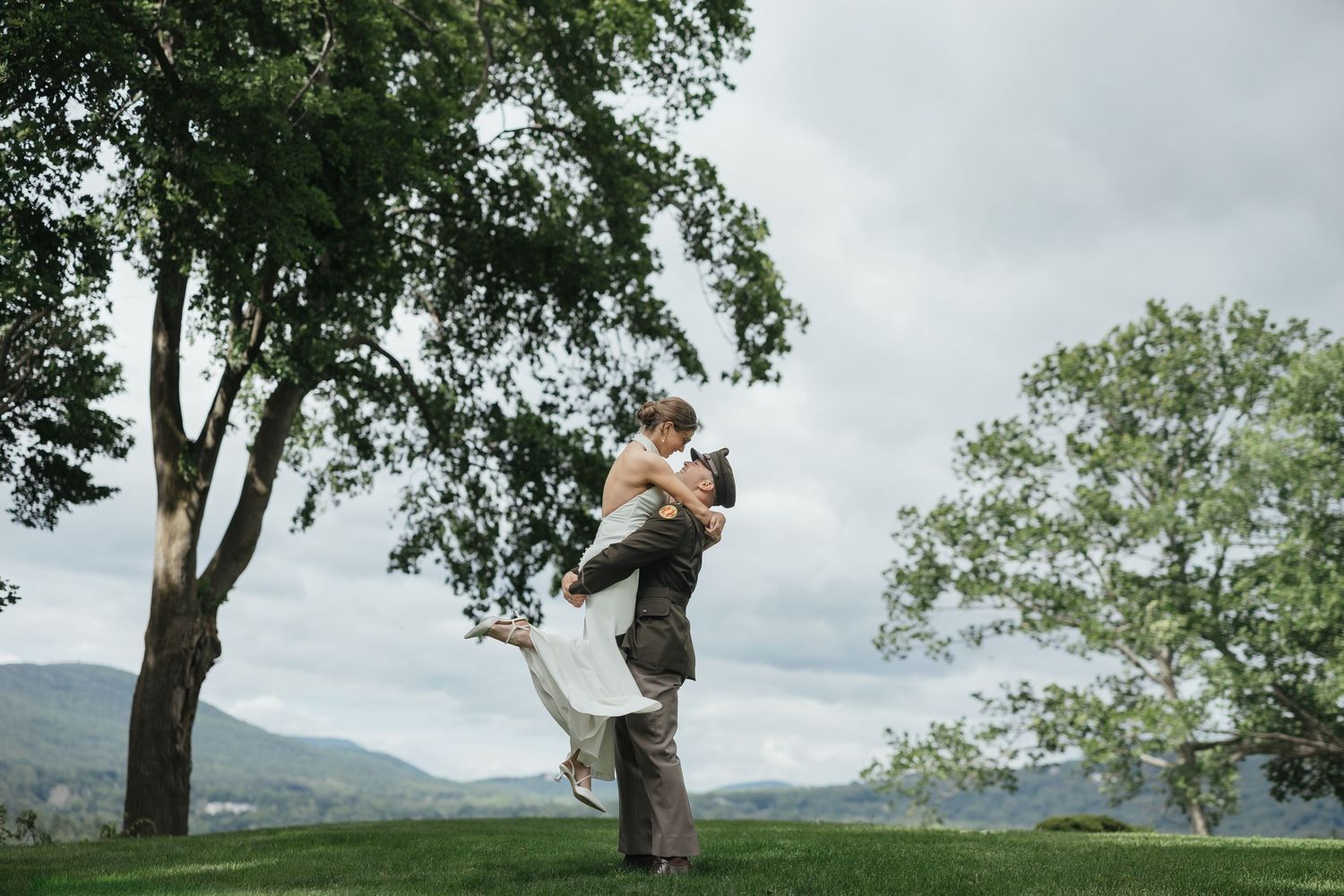 Groom lifting bride in wedding attire on green hillside with trees and mountain backdrop.