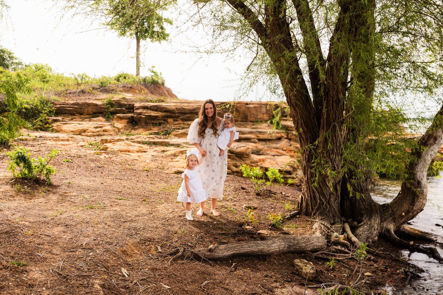 Mother and daughter in matching white floral dresses standing near a large tree with rocky cliffs in background.