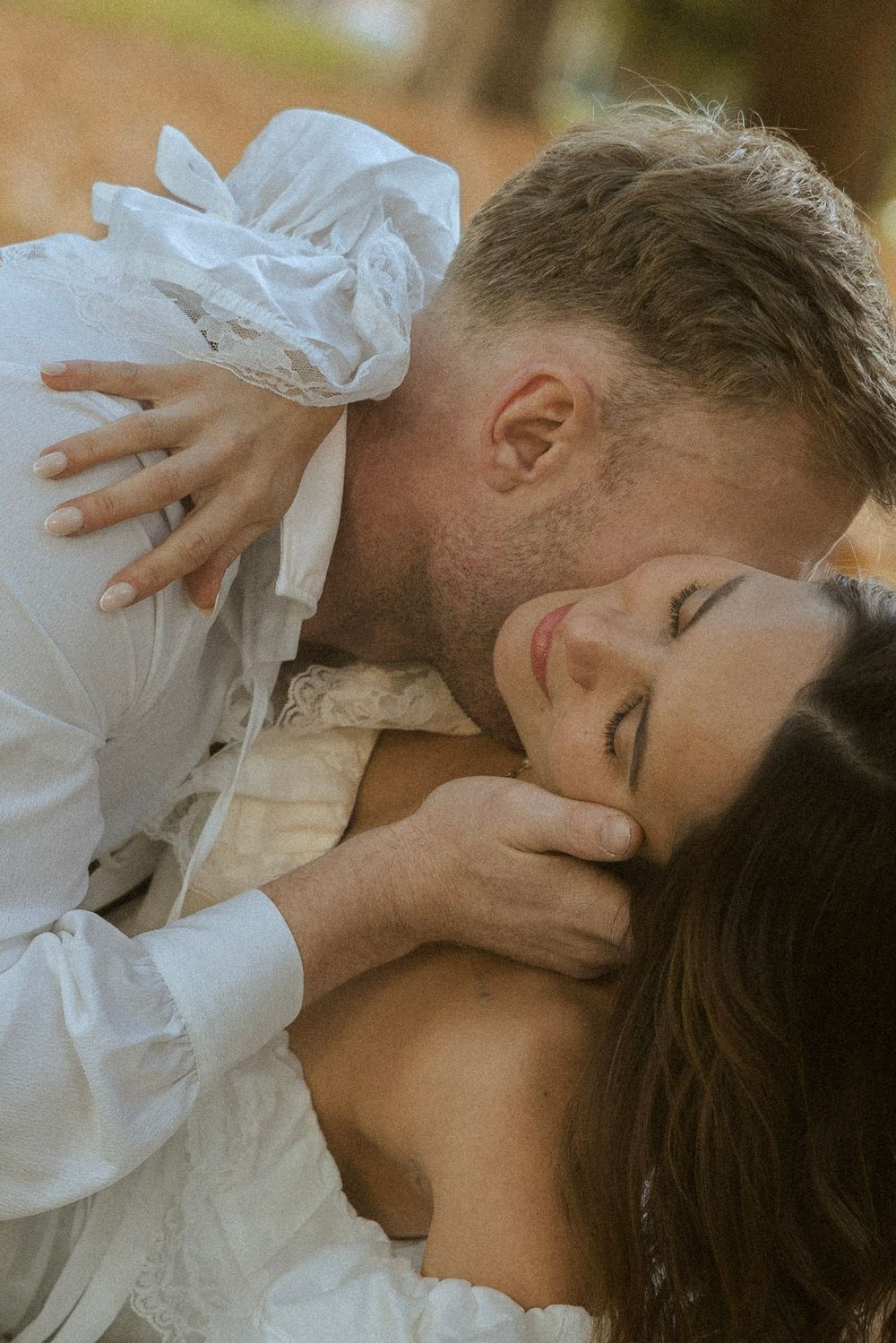 Groom tenderly kissing bride on cheek as she smiles with eyes closed in autumn setting.