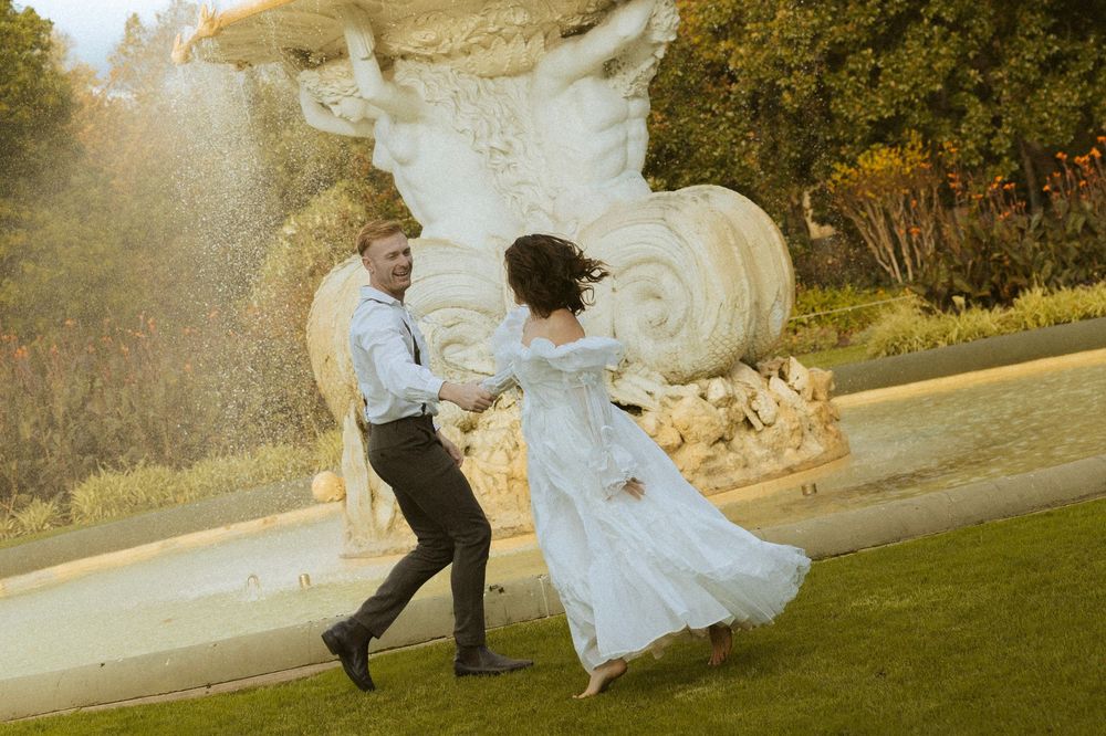 Bride and groom dancing joyfully near an ornate stone fountain in a lush garden.