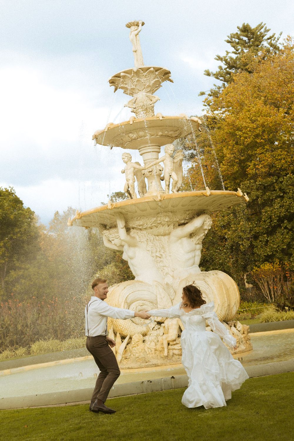 Bride and groom dancing joyfully in front of an ornate stone fountain in an autumn park.