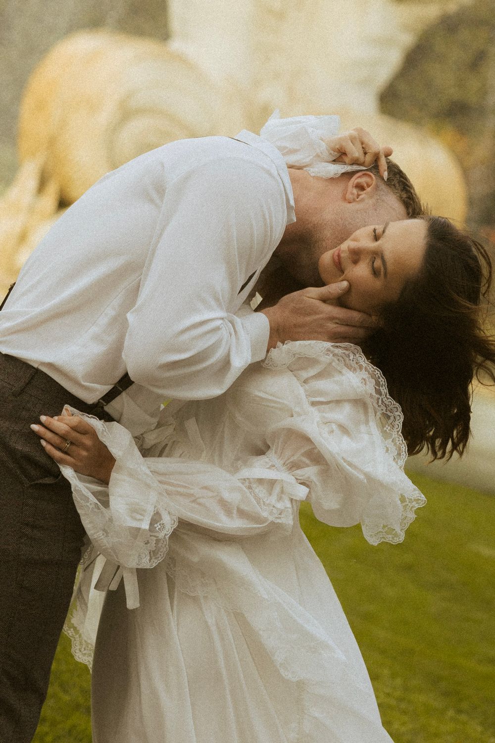 Groom kissing bride tenderly outdoors, both in elegant white wedding attire near a stone sculpture.