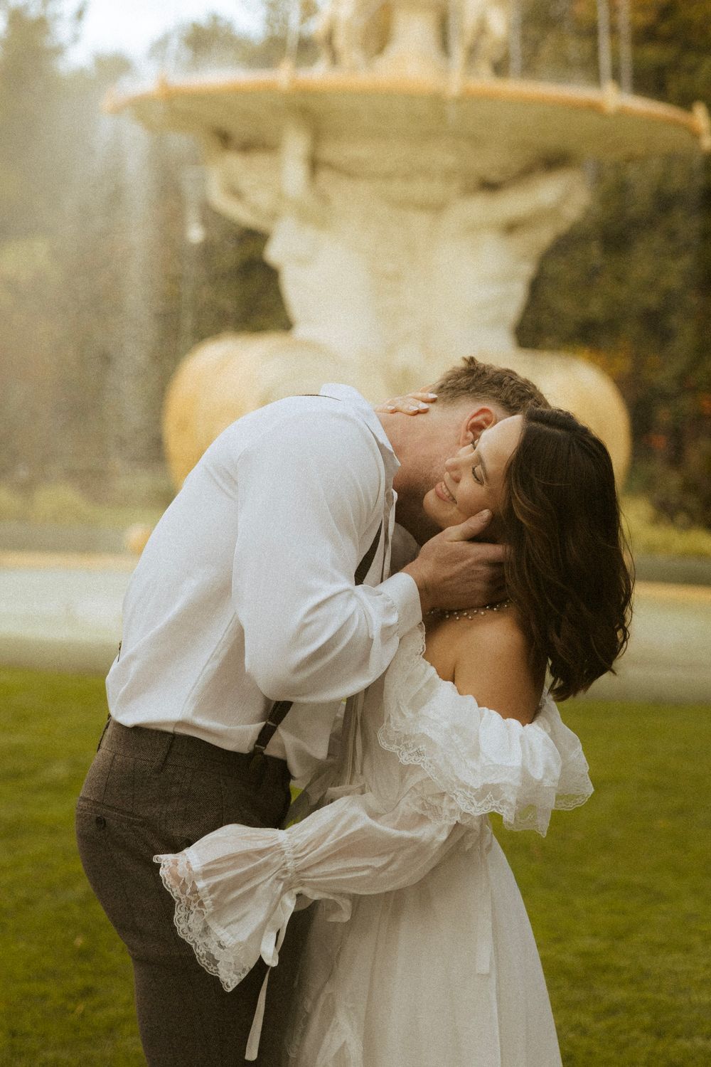 Groom kissing bride near ornate stone fountain in golden garden light.