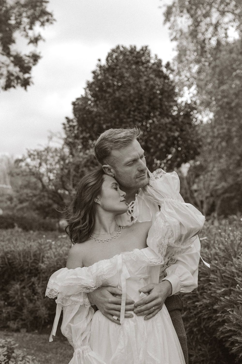 Black and white romantic portrait of couple embracing in garden, woman in off-shoulder white gown with puff sleeves.